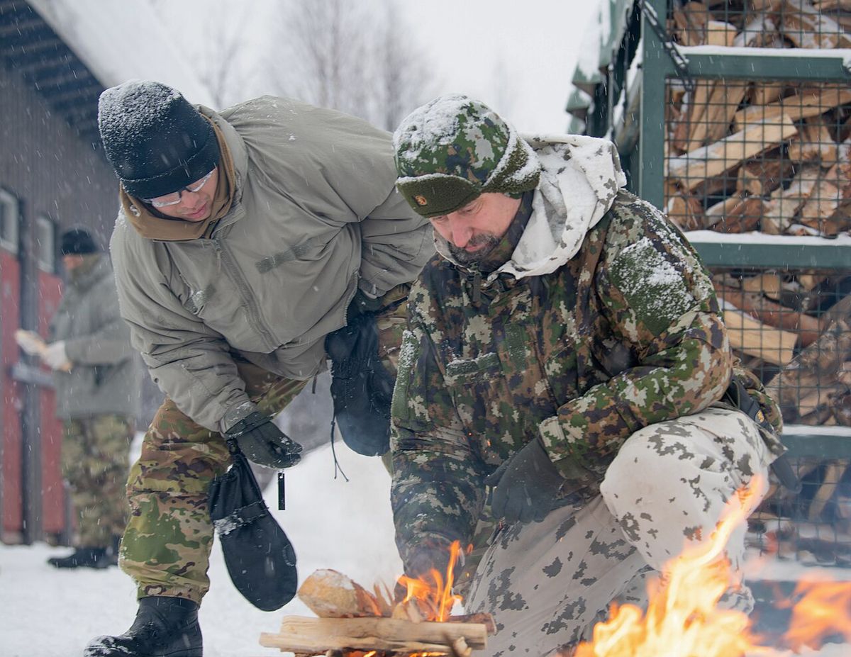 Two people in winter camouflage crouched over a small fire of twigs and bark