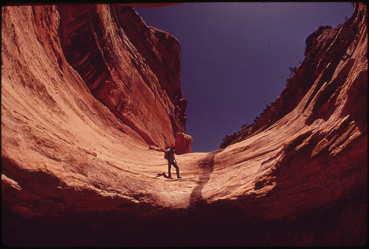 A hiker walking through a narrow red rock canyon toward distant light