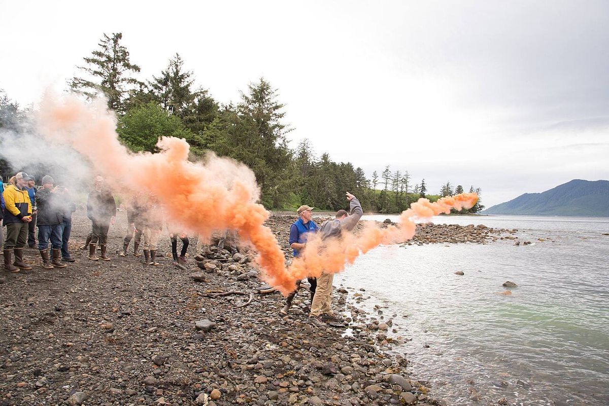 A person on a rocky shore releasing thick orange smoke toward the water