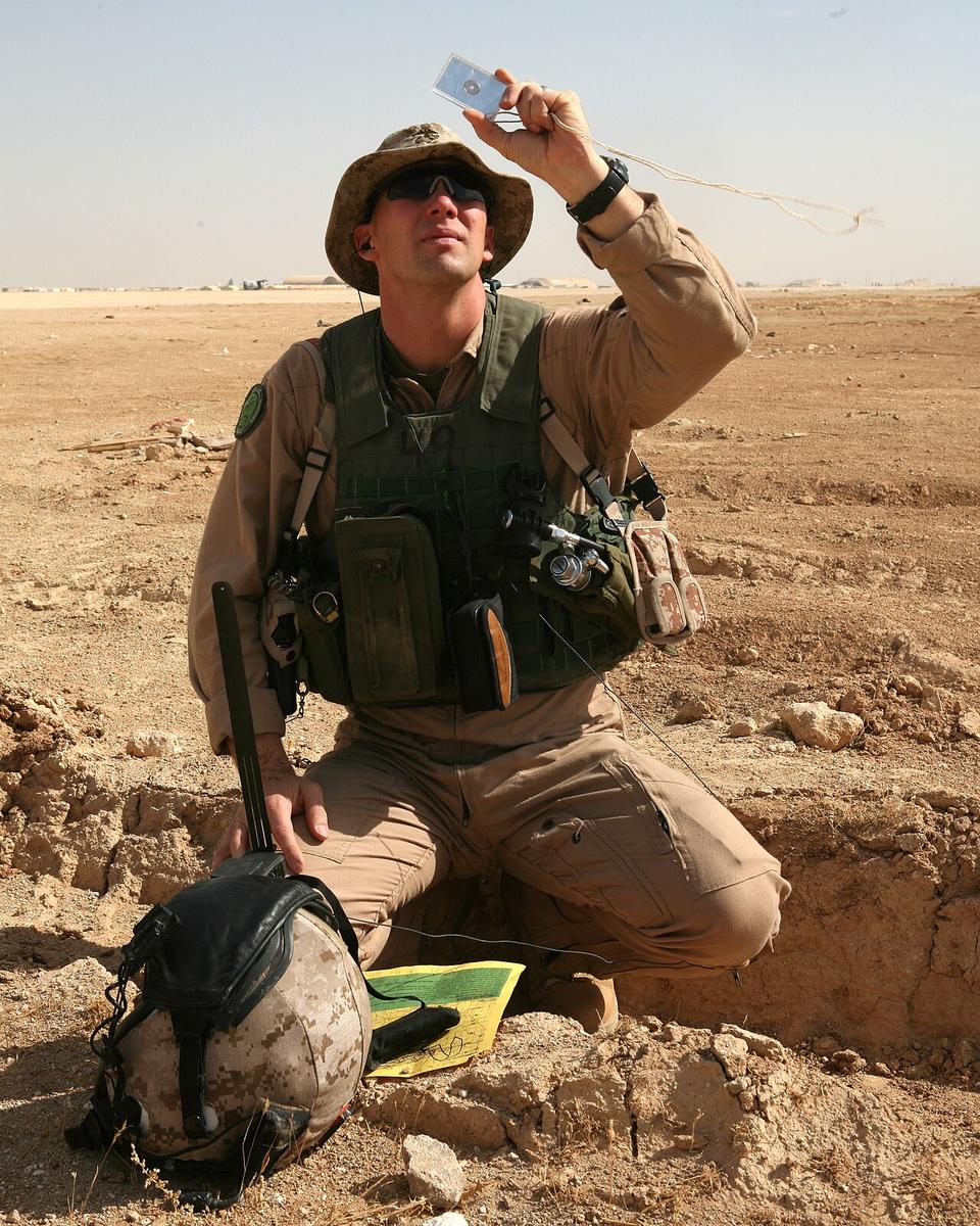 A Marine kneeling in a desert, holding a small mirror up toward the sky